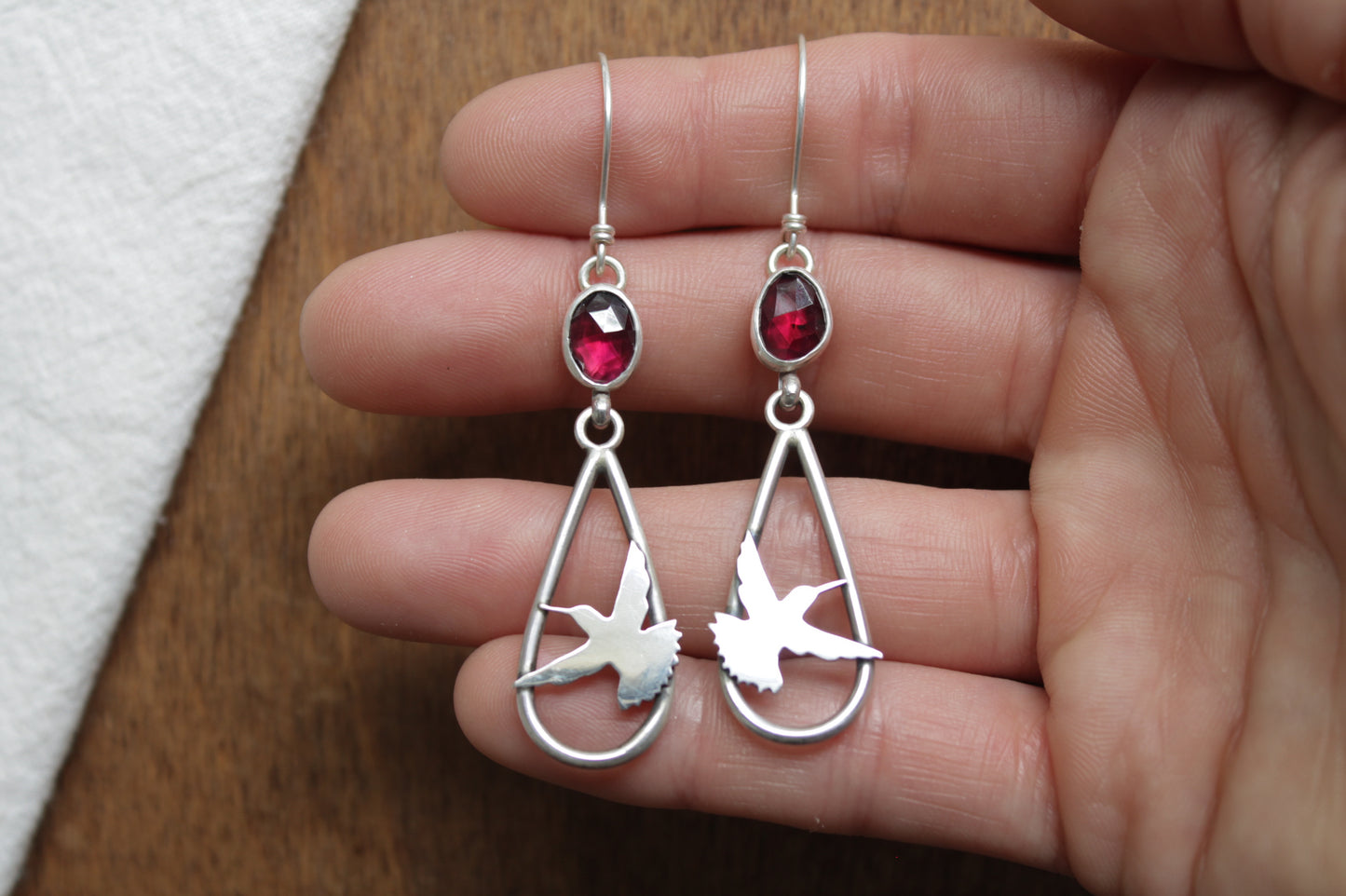 Silver earrings with teardrop design and red gemstones held in a hand on a wooden surface.