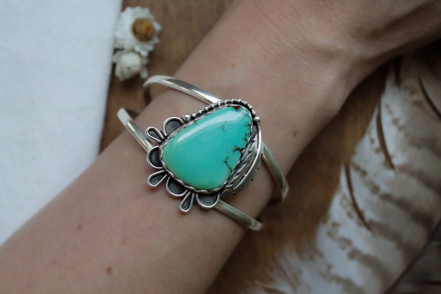 Turquoise Feather Statement Cuff bracelet showcasing a vibrant Iron Maiden Turquoise stone in a scalloped bezel. Romantic handmade details include intricate silver lace embellishments, a forged feather accent, and a satin finish. Adjustable cuff band made from responsibly sourced sterling silver. Ideal for nature-inspired style lovers.